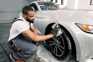 Car washing and detailing photo. African man worker in protective overalls and rubber gloves, washing car wheel rims of luxury white, on a car wash, using sponge and foam cleaning solution